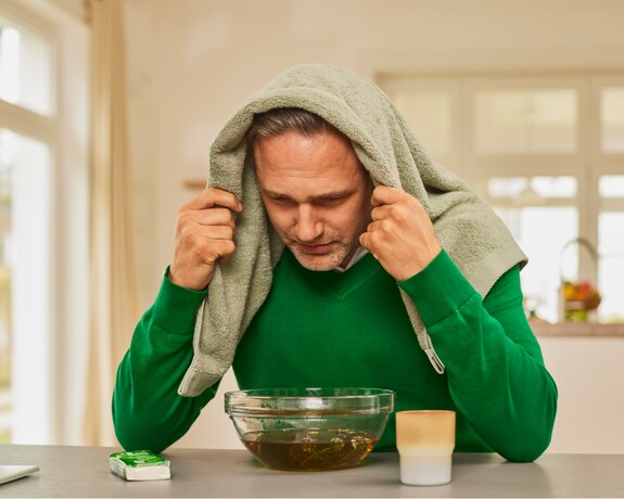 Mann in grünem Pullover mit einem Handtuch über dem Kopf beugt sich über eine Schüssel mit dampfendem Wasser.