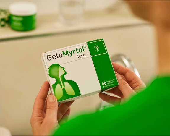 Gelomyrtol Forte – Vertrauen Sie auf bewährte Qualität für Ihre Erkältungslinderung. Hände halten eine Packung GeloMyrtol forte, die im Vordergrund scharf zu sehen ist, mit dem Logo und der grünen Markenfarbe deutlich sichtbar, im Hintergrund unscharf eine weitere Produktverpackung.