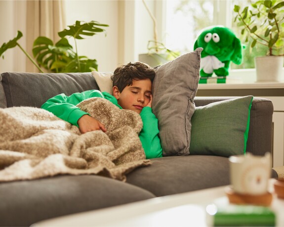 Junge in grünem Pullover liegt erschöpft auf einem Sofa unter einer kuscheligen Decke.