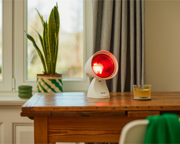 Eine Rotlichtlampe steht in einem gemütlichen Raum aktiviert auf einem Holztisch neben einer Sansevieria-Pflanze.