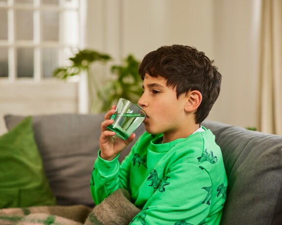 Kind mit Erkältung trinkt ein Glas Wasser auf dem Sofa.