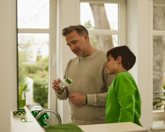 Die kindgerechten Myrtol® Kapseln lassen sich einfach schlucken und sind für Kinder ab 6 Jahren bestimmt. Vater und Sohn stehen nebeneinander im Badezimmer vor dem Spiegel und schauen sich die Verpackung vom schleimlösenden Präparat Myrtol® an.
