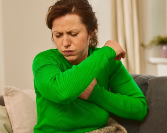 Gelomyrtol Forte – Linderung bei Erkältungssymptomen. Frau in grünem Pullover sitzt auf dem Sofa und hält sich den Arm, was auf Gliederschmerzen im Rahmen einer Erkältung hindeutet.
