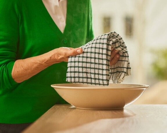 Gelomyrtol Forte – Traditionelle Hausmittel treffen auf bewährte Erkältungslinderung. Ältere Person in grünem Pullover bereitet als Hausmittel gegen Bronchitis einen warmen Wickel vor, indem sie ein kariertes Tuch über eine Schüssel mit heißem Wasser hält.