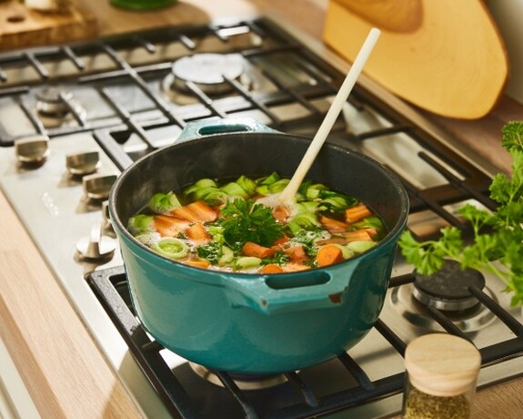 Frische Hühnersuppe köchelt auf einem Herd in einem blauen Topf, umgeben von Zutaten und Küchenutensilien.