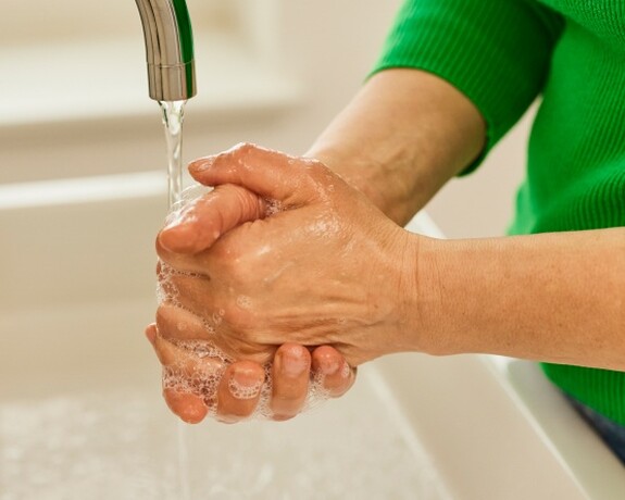 Handhygiene als wichtiger Schritt zur Vorbeugung vor Erkältungen. Person im grünen Pullover wäscht sorgfältig ihre Hände unter fließendem Wasser über einem Waschbecken.