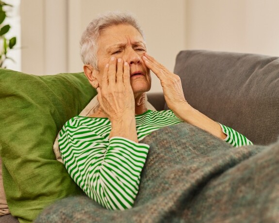 Gelomyrtol Forte - Effektive Unterstützung bei Sinusitis und Kieferhöhlenentzündung. Eine ältere Frau mit grauen Haaren, gekleidet in einem gestreiften grün-weißen Pullover, sitzt auf einem Sofa, hält sich die Handflächen an die Nebenhöhlen und scheint Schmerzen zu empfinden, möglicherweise aufgrund einer Kieferhöhlenentzündung.