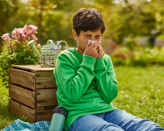 Ein Junge sitzt draußen auf einer Decke im Gras, gekleidet in einem grünen Pullover und blauen Jeans, und niest in ein Taschentuch.
