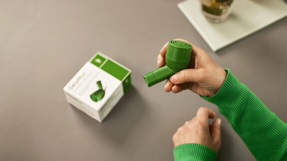 Eine Person hält das grüne Inhalationsgerät GeloMuc, mit einer  passenden Verpackung im Hintergrund, in der Hand.