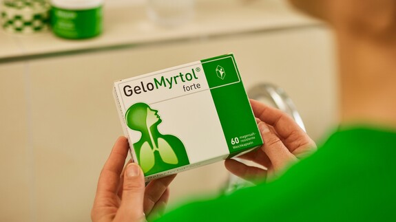 Hände halten eine Packung GeloMyrtol forte, die im Vordergrund scharf zu sehen ist, mit dem Logo und der grünen Markenfarbe deutlich sichtbar, im Hintergrund unscharf eine weitere Produktverpackung.